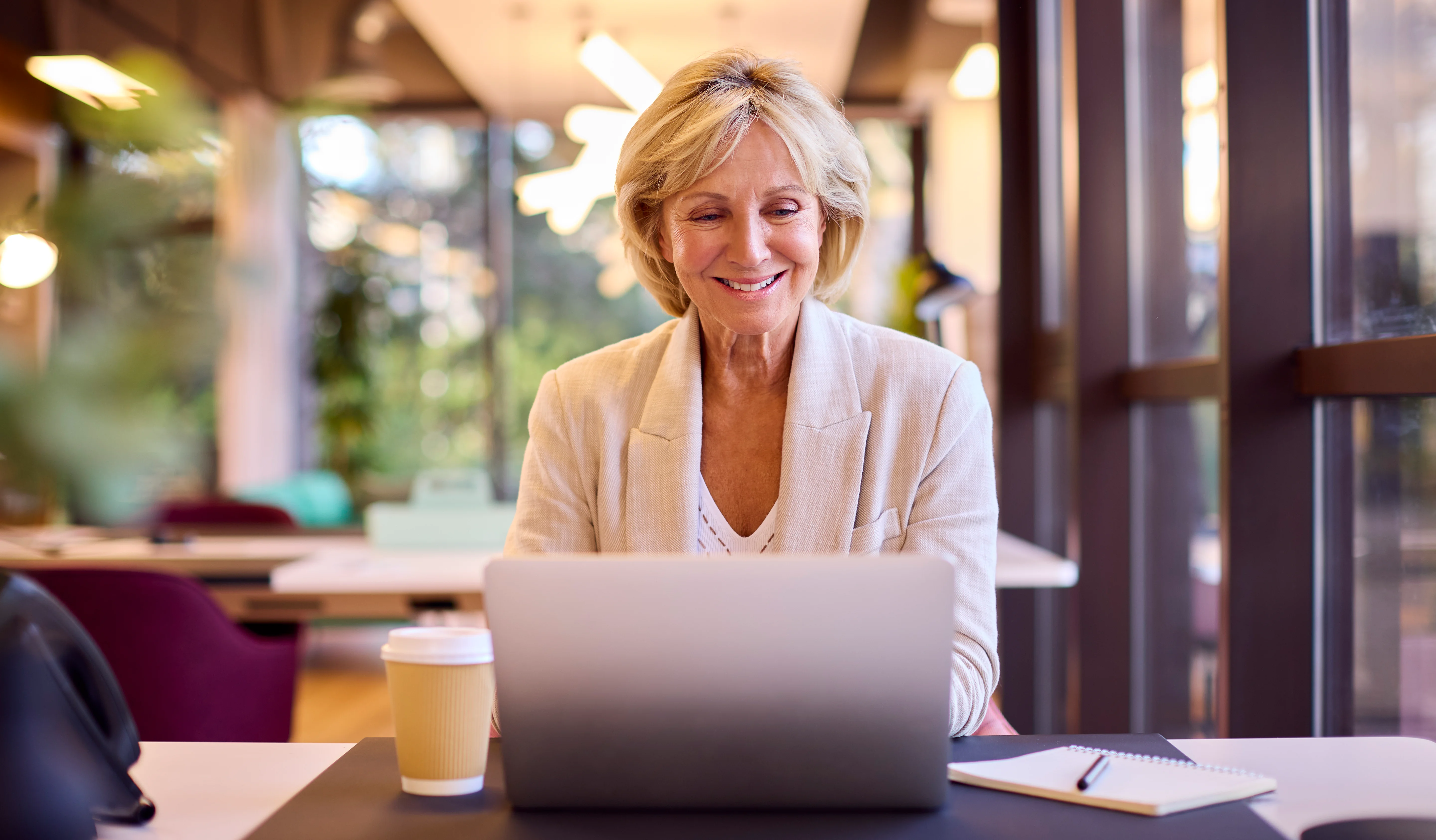 A businesswoman working on a laptop at a desk in an office with a takeaway drink.