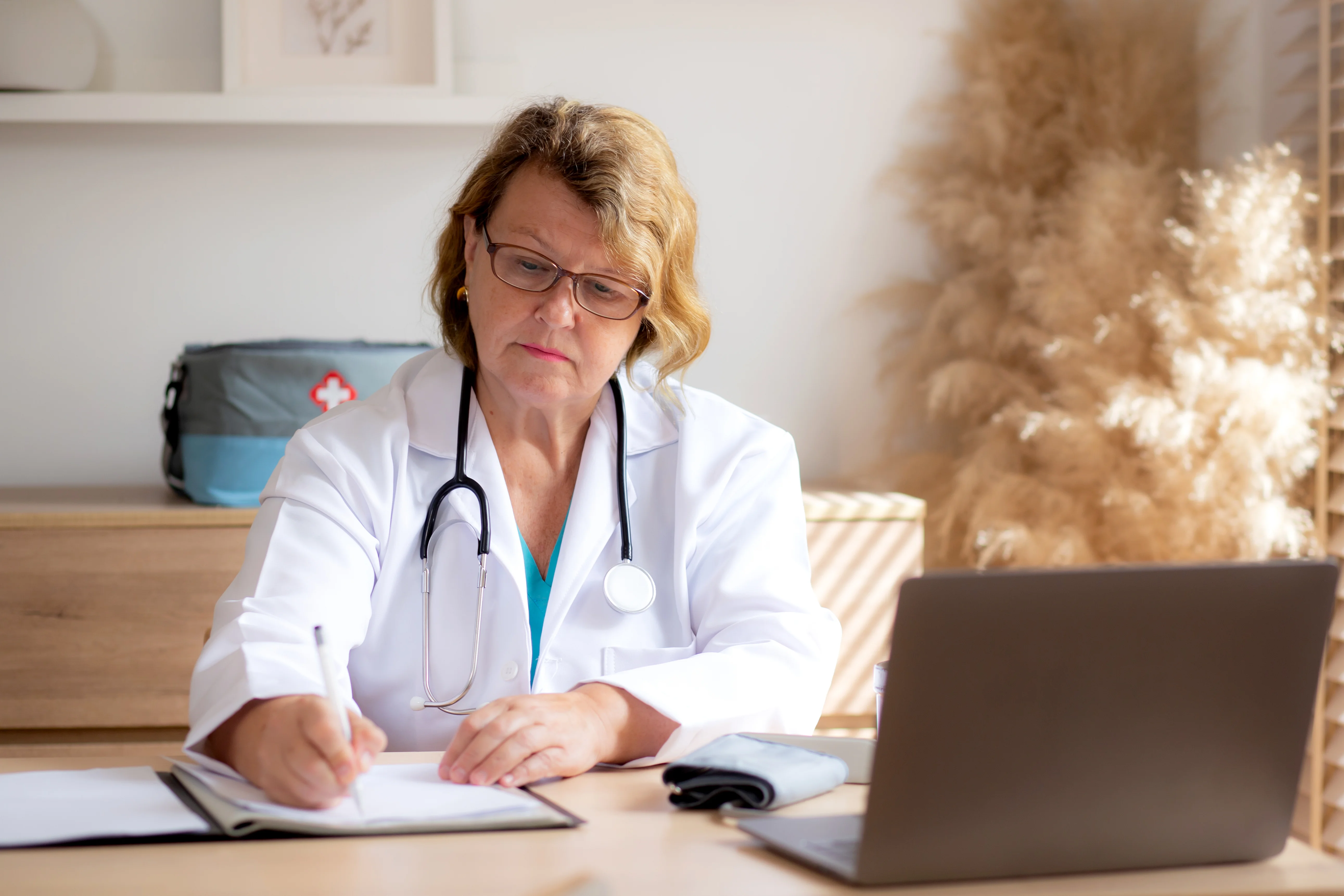 A doctor in a white coat with a stethoscope writing notes of a patient at a desk in a modern medical office. A laptop also rests on the desk.