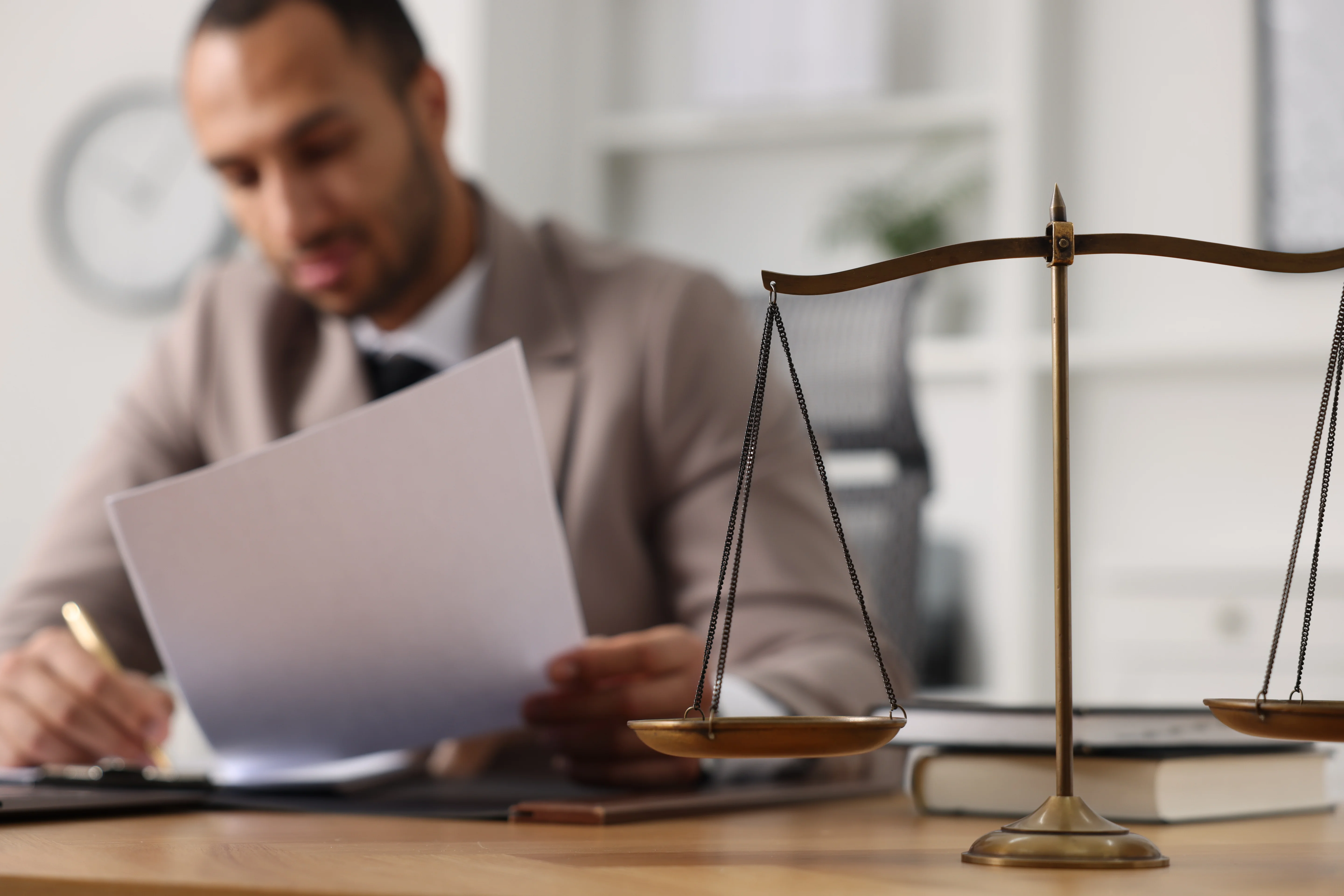 The scales of justice on a table in an office, with a lawyer, blurred, in the background working with a document at the table.